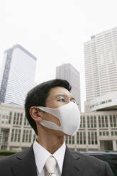 Portrait businessman in face mask below city highrise buildings - FSIF05694