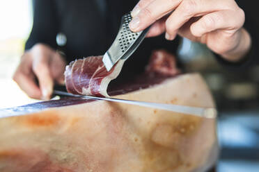 Young female chef cutting slice of ham with kitchen knife - JAQF00533