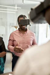 Lächelnder Geschäftsmann gestikuliert beim Betrachten der virtuellen Realität durch einen Simulator in einem Coworking-Büro - ZEDF04111