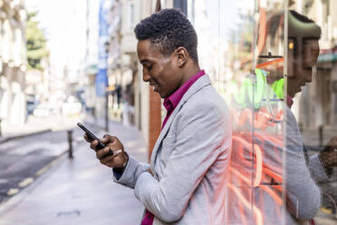 Männlicher Unternehmer, der ein Smartphone vor einer Glasscheibe benutzt - DLTSF01794
