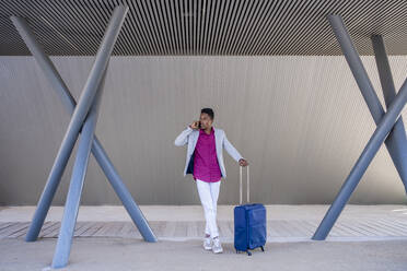 Businessman with suitcase looking away while talking on mobile phone under structure - DLTSF01798