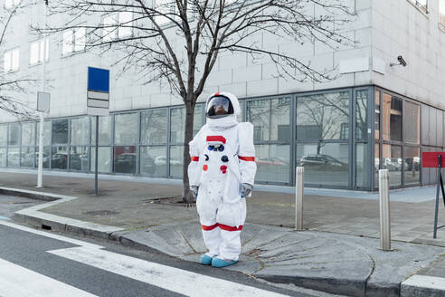 Junge Astronautin im Raumanzug steht in der Nähe eines Zebrastreifens vor einem Gebäude in der Stadt - MEUF02393