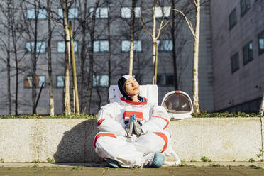 Female astronaut in space suit sitting with hands clasped by retaining wall during sunny day - MEUF02428