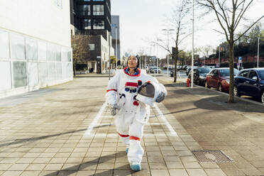 Smiling female astronaut holding space helmet while walking on footpath in city - MEUF02441