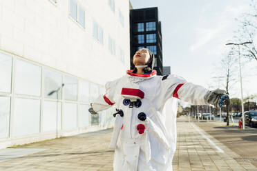 Young female astronaut with arms outstretched standing on footpath in city - MEUF02442