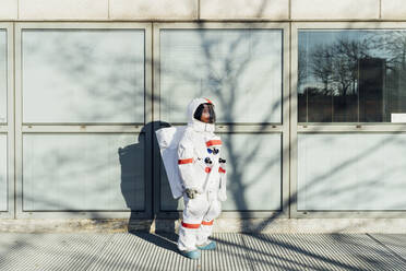 Junge Astronautin im Raumanzug steht an einem Glasfenster in der Stadt an einem sonnigen Tag - MEUF02443