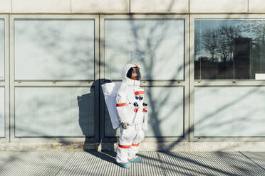 Young female astronaut in space suit standing by glass window in city during sunny day - MEUF02443