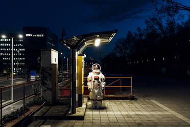 Woman astronaut standing under illuminated light at telephone booth in city during night - MEUF02509