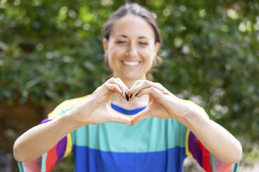 Smiling woman gesturing heart shape in garden - WPEF04307