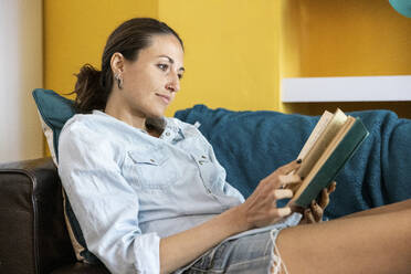 Woman reading book while sitting on sofa at home - WPEF04324
