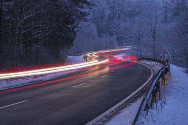 Deutschland, Bayern, Spessart, Lichterweg auf kurvenreicher Straße durch majestätischen Winterwald in der Abenddämmerung - RUEF03254
