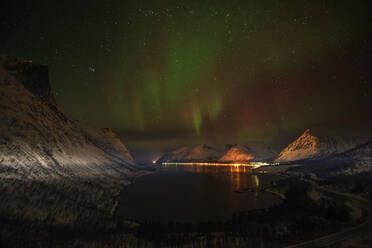 Norwegen, Tromso, Bergsbotn, Blick auf die Aurora Borealis über dem beleuchteten Fjorddorf auf der Insel Senja - RUEF03278