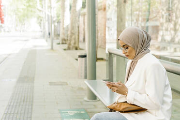 Young woman using smart phone while sitting on bench at station - XLGF01569