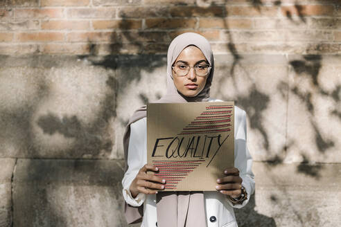 Junge Frau hält Plakat mit Gleichstellungstext vor einer Mauer an einem sonnigen Tag - XLGF01573