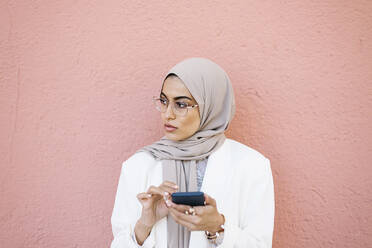 Schöne Frau, die ihr Smartphone vor einer rosa Wand hält und wegschaut - XLGF01590