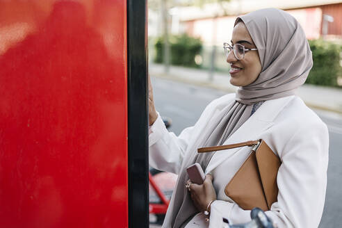 Lächelnde Frau mit Handtasche und Smartphone in der Hand bei der Benutzung eines Selbstbedienungsautomaten an der Straße - XLGF01591