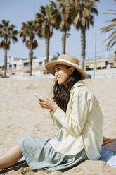 Smiling woman looking away while holding smart phone on beach during sunny day - AFVF08662