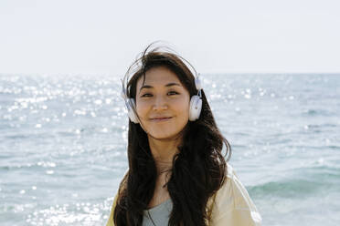 Beautiful woman with in-ear headphones standing on beach during sunny day - AFVF08681