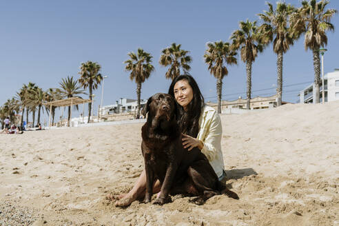 Frau, die ihren Hund streichelt, während sie an einem sonnigen Tag im Sand am Strand sitzt - AFVF08687