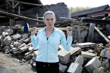 Selbstbewusste Frau, die einen Hammer hält, während sie vor einem zerstörten Haus steht - FLLF00592