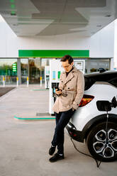 Männlicher Unternehmer in stilvollem Outfit, der an einer Tankstelle steht und auf seinem Mobiltelefon surft, während er in der Nähe eines Autos mit Zapfpistole steht - ADSF23199