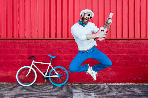 Playful verrückt bärtigen männlichen Radfahrer springen mit offenem Mund, während Ukulele Instrument in der Nähe von Fahrrad auf der Straße in der Nähe von roten Hintergrund spielen - ADSF23231