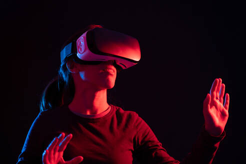Unerkennbare junge Frau in Freizeitkleidung und VR-Headset macht neue Erfahrungen und berührt virtuelle Objekte in einem Raum mit bunter Projektorbeleuchtung - ADSF23243
