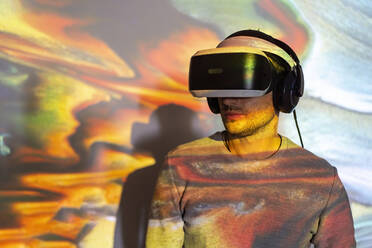 Unrecognizable young male in casual wear and VR headset getting new experience and touching virtual object in room with colorful projector illumination - ADSF23247