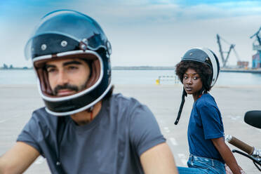 Stilvolles junges multiethnisches Paar in Freizeitkleidung und Helmen, das sich nach einer Motorradfahrt auf der Promenade am Meer ausruht - ADSF23262
