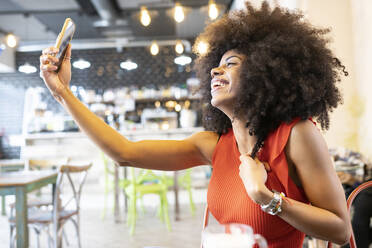 Fröhliche afroamerikanische Frau, die in einem Café sitzt und sich mit ihrem Smartphone fotografiert, während sie das Wochenende genießt - ADSF23272