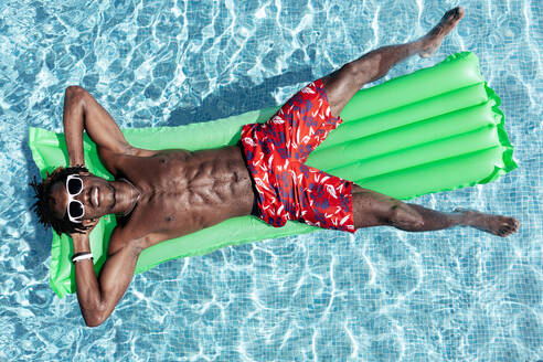 Sorgloser afroamerikanischer Mann mit nacktem Oberkörper in Shorts und Sonnenbrille liegt auf einer aufblasbaren Matratze im Schwimmbad und genießt den sonnigen Tag im Sommerurlaub - ADSF23278