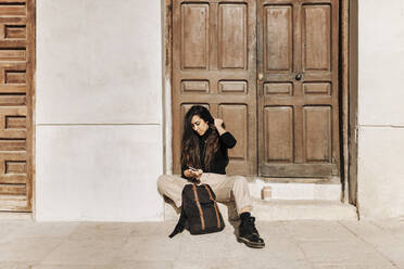 Young woman using mobile phone while sitting with backpack at doorway - MRRF01037