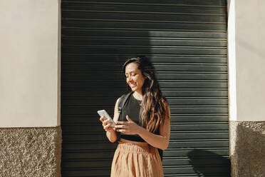 Lächelnde Touristin, die ein Smartphone benutzt, während sie vor einem Fensterladen steht - MRRF01045