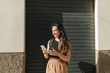 Smiling female tourist using smart phone while standing in front of shutter - MRRF01045