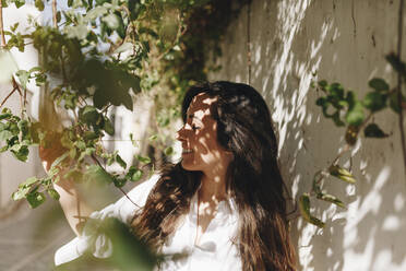 Woman smiling while touching plant by surrounding wall - MRRF01071