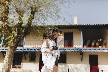 Woman with hand in hair holding camera at village - MRRF01085