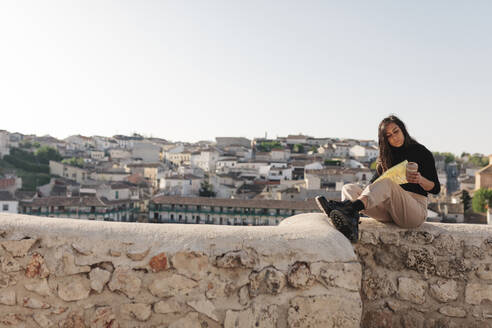 Frau prüft Karte, während sie an einem sonnigen Tag auf einer Stützmauer sitzt - MRRF01091