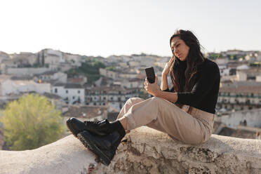 Weibliche Touristin, die ein Selfie mit ihrem Handy macht, während sie auf einer Stützmauer sitzt - MRRF01092