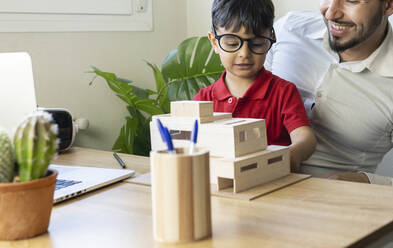 Junge mit Brille betrachtet ein Hausmodell, während er mit seinem Vater zu Hause sitzt - JCCMF02012