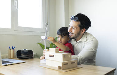 Männlicher Architekt lächelt, während sein Sohn mit dem Modell einer Windkraftanlage im heimischen Büro spielt - JCCMF02021