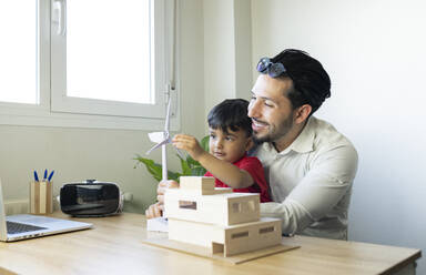 Male architect smiling while son playing with wind turbine model at home office - JCCMF02021