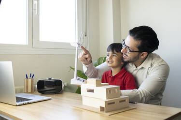 Male architect pointing at wind turbine while sitting with son at home office - JCCMF02022