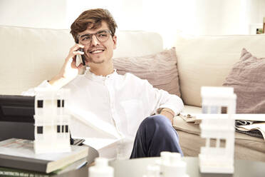 Lächelnder männlicher Architekt, der auf dem Sofa sitzend mit seinem Handy telefoniert - UKOF00167