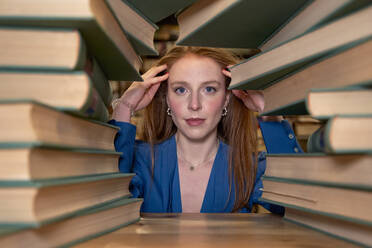 Schöne Frau mit Hand im Haar durch Stapel von Büchern in der Bibliothek gesehen - VEGF04337