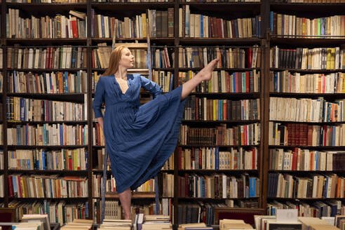 Junge Frau in blauem Kleid balanciert auf einem Bein auf einer Leiter in einer Bibliothek - VEGF04359