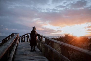 Seitenansicht einer älteren weiblichen Reisenden in Freizeitkleidung, die auf einem Holzsteg am Sandstrand steht und das Meer bei Sonnenuntergang genießt - ADSF23297