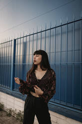 Stylish young ethnic female with long dark hair in trendy dress standing against blue wall on street and looking away thoughtfully - ADSF23304