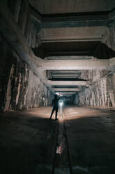 Back view of anonymous person with glowing flashlight in hand standing in abandoned tunnel with shabby walls - ADSF23326