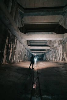 Rückenansicht einer anonymen Person mit einer leuchtenden Taschenlampe in der Hand, die in einem verlassenen Tunnel mit schäbigen Wänden steht - ADSF23326