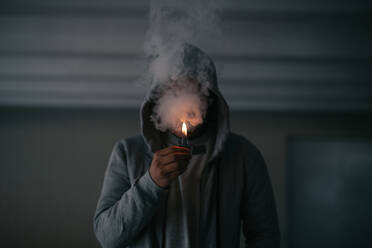 Unrecognizable male in hooded jacket smoking cigarette while standing in dark room with burning lighter in hand - ADSF23327